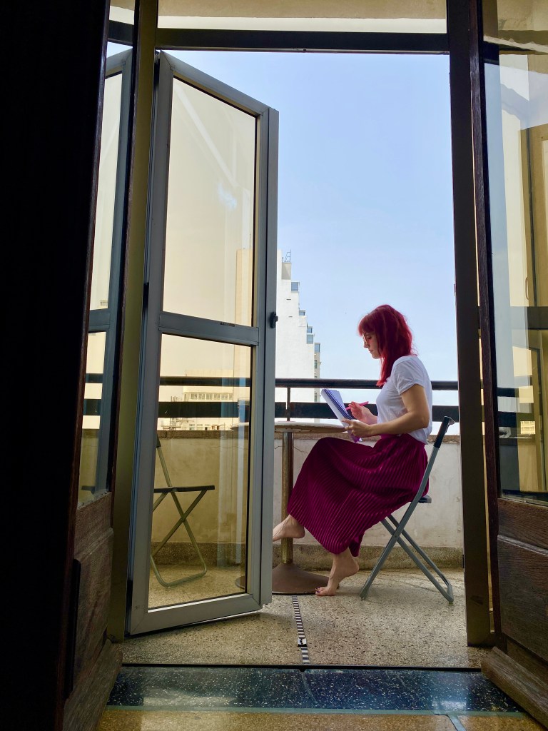 A red haired, white woman in a white top and pink skirt sits reading on a balcony.